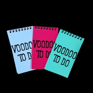 Puede incluir: Tres blocs de notas con espiral en azul claro, rosa intenso y turquesa. Cada bloc de notas tiene las palabras "VOODOO TO DO" impresas en negro. Los blocs de notas están sobre un fondo negro.