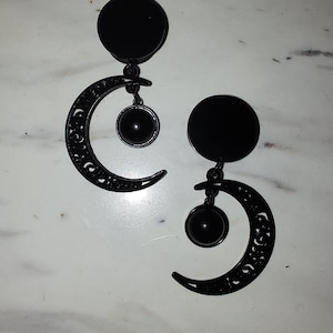 May include: A pair of black earrings featuring a circular stud, a crescent moon design with intricate cutouts, and a dangling black sphere. The earrings have a gothic or celestial theme.