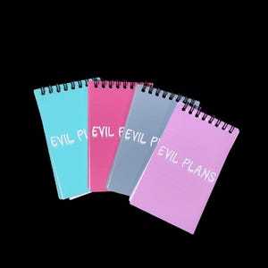 Puede incluir: Cuatro pequeños cuadernos de espiral con el texto "Evil Plans" escrito en blanco en cada portada. Los cuadernos son de diferentes colores: turquesa, rosa, gris y rosa claro.