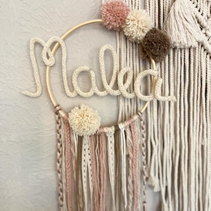 Pode incluir: Uma decoração de parede circular em madeira com o nome Malea formado por um cordão de malha cor de creme. O aro é decorado com pompons macios em tons de rosa, creme e castanho, com fios pendentes de renda, lã e cordão entrançado.