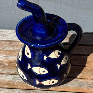 May include: A dark blue ceramic pitcher with a distinctive spout and handle. The pitcher is decorated with white fish designs and red accents. The design has a nautical theme. The pitcher is likely used for serving beverages.