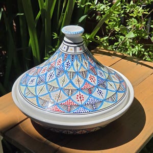 May include: A ceramic tagine featuring a conical lid with a white base and a detailed, repeating geometric pattern in shades of blue, red, and yellow. The lid has a small, circular opening at the top with blue and white striped detailing.