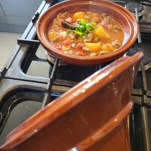 May include: A close-up of a brown tagine pot filled with a stew of beef, potatoes, tomatoes, and herbs. The pot is on a black stovetop with a matching brown lid.