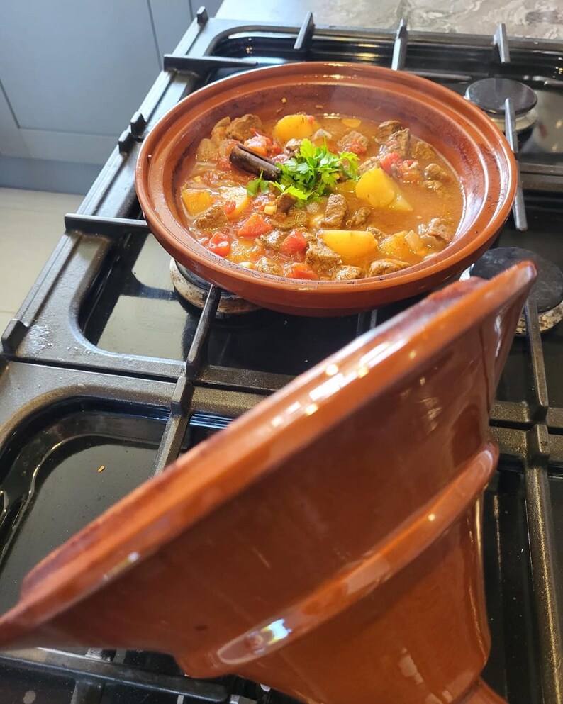 May include: A close-up of a brown tagine pot filled with a stew of beef, potatoes, tomatoes, and herbs. The pot is on a black stovetop with a matching brown lid.