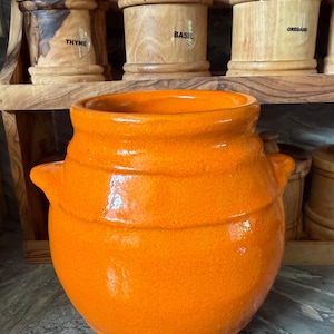 May include: A bright orange ceramic jar with a wide mouth, a rounded body, and two small side handles. The jar features a glossy, textured finish and sits in front of a wooden shelf containing three wooden canisters labelled Thyme, Basil, and Oregano.
