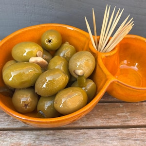 May include: A bright orange ceramic olive dish containing green olives and toothpicks. The dish is divided into two sections, one for olives and the other for toothpicks. The olives are shiny and appear to be stuffed.