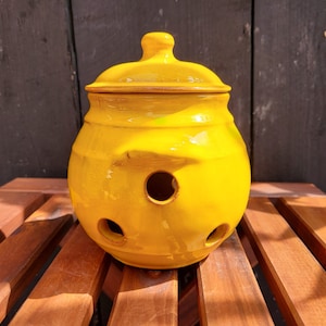 May include: A yellow ceramic jar with a lid and holes for ventilation. The jar is sitting on a wooden surface.