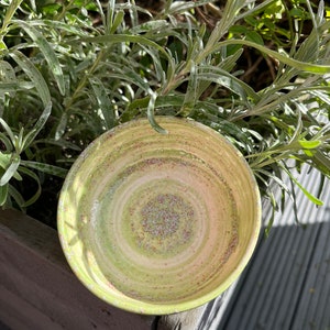 May include: A green ceramic bowl with a white and pink swirl design and a speckled finish. The bowl is sitting on a wooden surface.