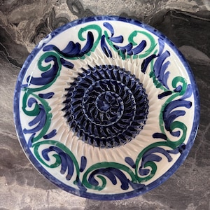 May include: A white ceramic grater plate with a blue and green floral design around the rim. The centre features a textured, dark blue grating surface. The plate is designed for grating ingredients.