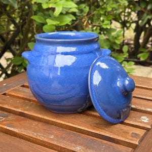 May include: A vibrant cobalt blue ceramic jar with a lid, placed on a wooden table. The jar has two small handles and a rounded form. The lid is partially open. The background shows green foliage.