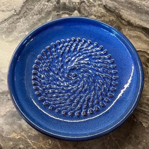 Puede incluir: Un plato rallador de cer&aacute;mica circular con un esmalte azul cobalto vibrante. El centro presenta una textura en relieve con patr&oacute;n de espiral dise&ntilde;ada para rallar alimentos, rodeada por un borde ancho y liso. El acabado tiene un brillo sutil.