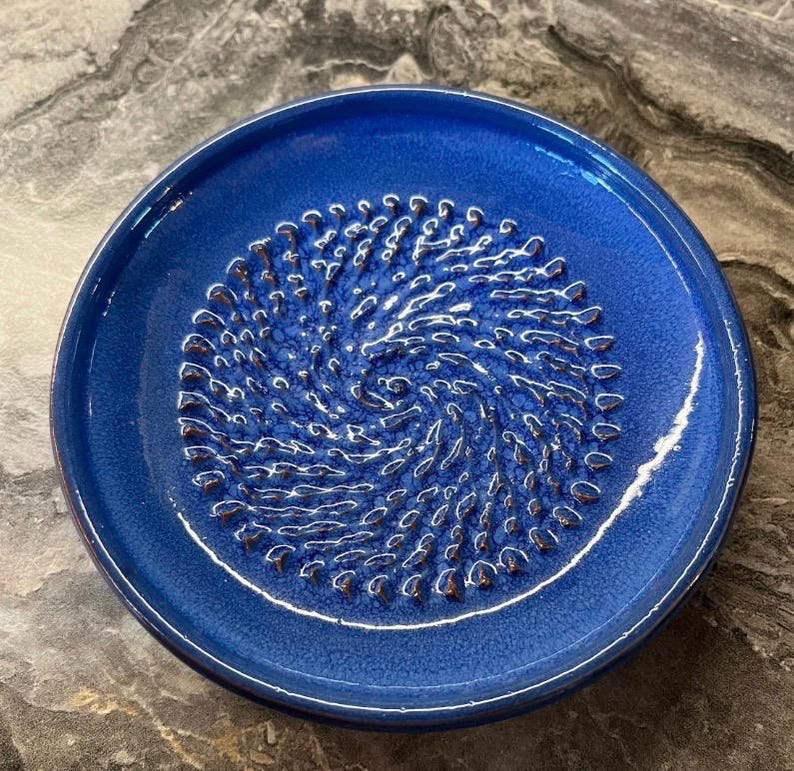 Puede incluir: Un plato rallador de cer&aacute;mica circular con un esmalte azul cobalto vibrante. El centro presenta una textura en relieve con patr&oacute;n de espiral dise&ntilde;ada para rallar alimentos, rodeada por un borde ancho y liso. El acabado tiene un brillo sutil.