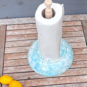 May include: A white paper towel roll on a decorative holder. The holder has a light blue and white marbled design. The wooden dowel is visible inside the paper towel roll. The holder is on a wooden table.