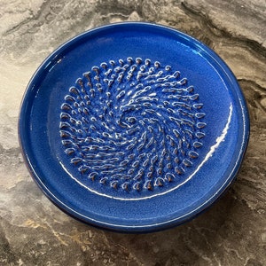 May include: A blue ceramic garlic grater with a textured surface. The grater is round and has a raised centre.