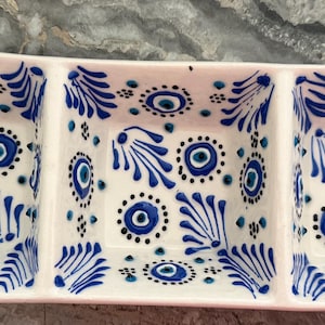 May include: A rectangular ceramic serving dish with three square compartments. The white surface is decorated with a hand-painted pattern of blue and black circular eye motifs and stylised blue leaf designs across all three sections.