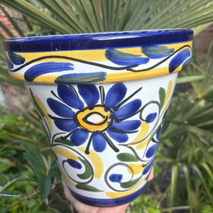 May include: A hand-painted ceramic flower pot with a blue, yellow, and white floral design. The pot features a large blue flower on the front and decorative swirls. The rim is blue with yellow and blue accents. The pot is approximately 25 cm tall.