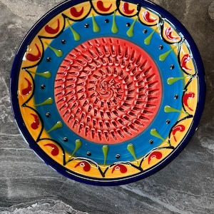 May include: A colourful ceramic plate with a red, blue and yellow design. The centre of the plate has a grater pattern.