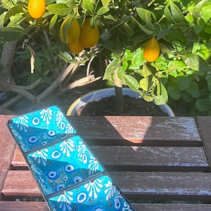 May include: A rectangular, turquoise serving dish with three compartments. Each compartment is decorated with white floral and blue eye designs. The dish is on a wooden table, with a small lemon tree in the background.