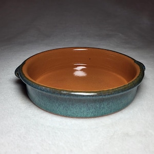 May include: A handmade, round ceramic baking dish with a teal exterior and a warm brown interior. The dish has two small handles and a glossy finish. This stoneware dish is perfect for baking and serving.