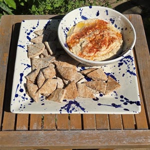 May include: A white square platter with blue paint splatters holds pita chips and a bowl of hummus sprinkled with paprika. The bowl is white with a blue rim. The food is on a wooden table.