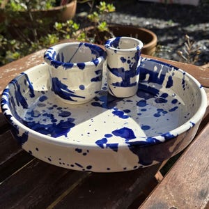 May include: A ceramic set consisting of a shallow round tray, a small wide cup, and a tall narrow cylinder, all featuring a glossy off-white base glaze decorated with a vibrant cobalt blue paint splatter pattern across the entire surface.