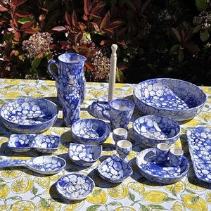 May include: A selection of blue and white ceramic tableware, including bowls, plates, a jug, and cups. The items have a speckled design and are displayed on a yellow and white patterned tablecloth. The set includes a serving spoon and a rectangular platter.