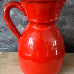 May include: A bright red ceramic jug with a curved handle and a spout. The jug has a glossy finish and a rounded body. The handle is thick and sturdy, and the spout is designed for easy pouring. Suitable for serving beverages.