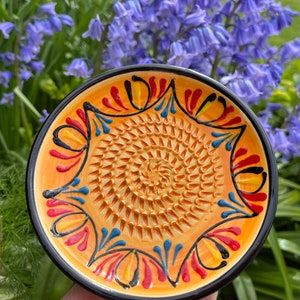 May include: A ceramic garlic press with a yellow, orange, red, blue and black design. The grater is in the centre of the plate.