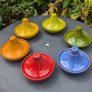 May include: Six colourful ceramic serving dishes with a rounded, dome-shaped lid. The dishes are arranged in a circle on a grey surface. The colours of the dishes are yellow, orange, red, blue, green, and a lighter shade of yellow.