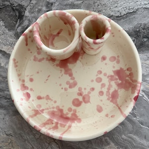 May include: A cream-coloured ceramic set consisting of a large round shallow bowl, a medium-sized cylindrical cup, and a small cylindrical cup. Each piece features a hand-painted splatter pattern in a dusty rose pink colour across the smooth surface.