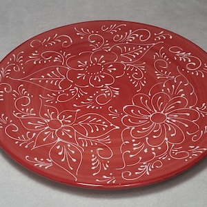 May include: A red ceramic plate with a white floral design. The plate has a slightly raised rim and a smooth, glossy finish.