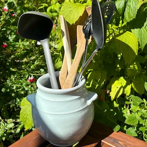 May include: A white ceramic utensil holder with a rounded shape and two handles. The holder contains a variety of kitchen utensils, including a black ladle, a wooden spoon, a black slotted spoon, and a black wire whisk.