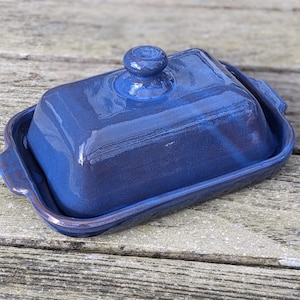 May include: A blue ceramic butter dish with a lid. The dish has a smooth, glossy finish and a slightly rounded shape.