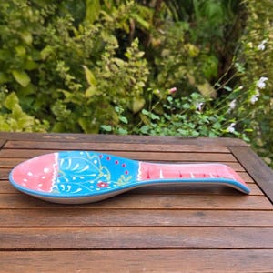 May include: A ceramic spoon rest with a pink and blue floral design. The spoon rest has a curved shape and is decorated with a white floral pattern and red accents. The edges are outlined in blue.