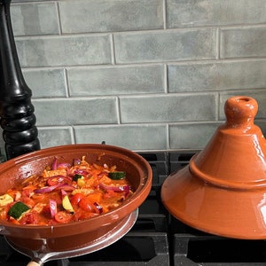 May include: A red clay tagine pot with a lid on a stovetop. The pot is filled with a stew of vegetables and meat.