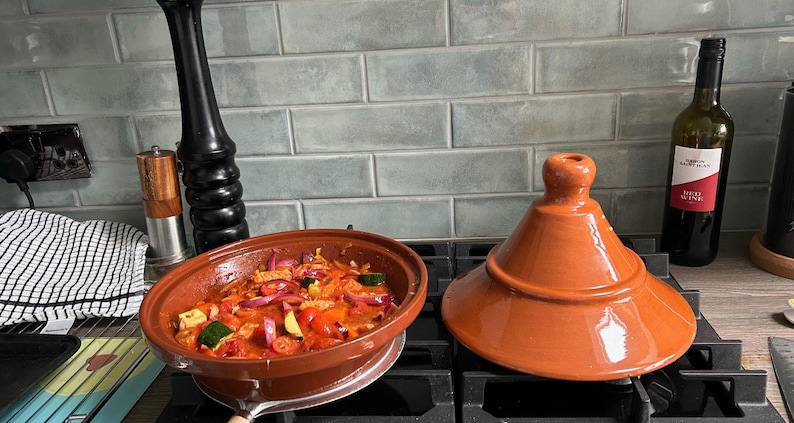 May include: A red clay tagine pot with a lid on a stovetop. The pot is filled with a stew of vegetables and meat.