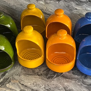 May include: A selection of vibrantly coloured ceramic salt pig bowls in yellow, green, orange, blue, and red. Each bowl features a rounded top with a small knob and a wide opening. The bowls are displayed on a textured surface.