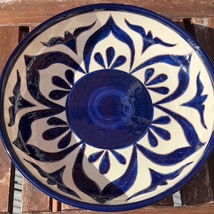 May include: A round ceramic bowl featuring a deep blue circular centre surrounded by a symmetrical pattern of dark blue floral and leaf-like motifs painted against a cream-coloured background. The bowl has a smooth, glossy finish throughout.