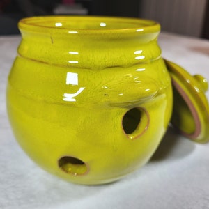 May include: A bright yellow ceramic jar with a lid. The jar has a hole in the side and is designed for storing garlic or onions.
