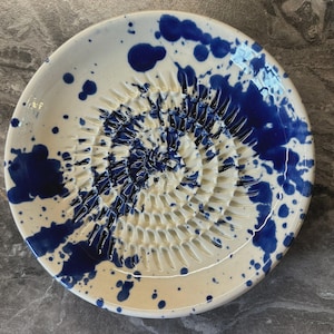 Puede incluir: Un plato rallador redondo de cer&aacute;mica con base blanca y un patr&oacute;n de salpicaduras azul vibrante. El centro del plato tiene una textura elevada en forma de espiral para rallar alimentos, con esmalte azul en las ranuras.