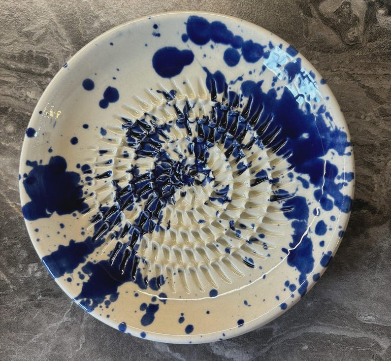 Puede incluir: Un plato rallador redondo de cer&aacute;mica con base blanca y un patr&oacute;n de salpicaduras azul vibrante. El centro del plato tiene una textura elevada en forma de espiral para rallar alimentos, con esmalte azul en las ranuras.