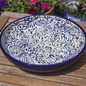 May include: A round ceramic serving bowl with a blue rim and intricate blue floral patterns on a cream background. The bowl is on a wooden table. Plants are visible in the background.