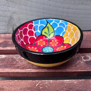May include: A small, round ceramic bowl with a black rim and a yellow base. The interior features a vibrant floral design with red, blue, yellow, and white colours. The bowl sits on a wooden surface.