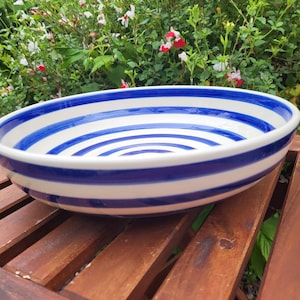May include: A large, round ceramic bowl with a white interior and exterior. The bowl features a bold, concentric design of alternating blue and white stripes. The bowl is placed on a wooden surface, with greenery in the background.