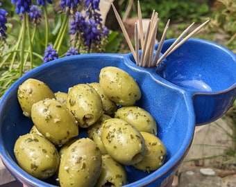 May include: A blue ceramic serving dish with two compartments. One compartment holds green olives with herbs, the other compartment is empty. The dish is on a table with a blurred background of green plants.