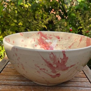 May include: A large, cream-coloured ceramic bowl with a wide rim and a rounded base. The bowl features striking pink splatters, creating a unique design. The bowl is placed on a wooden surface.