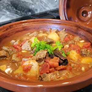May include: A close-up of a brown tagine pot filled with a stew of beef, potatoes, tomatoes, and onions. The stew is topped with fresh parsley and a cinnamon stick.