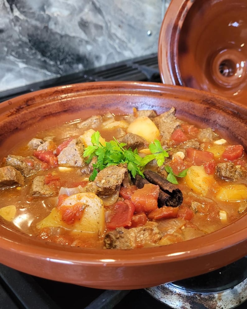 May include: A close-up of a brown tagine pot filled with a stew of beef, potatoes, tomatoes, and onions. The stew is topped with fresh parsley and a cinnamon stick.