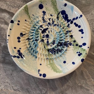 Puede incluir: Un plato rallador de cer&aacute;mica redondo con base color crema, que presenta una espiral central de dientes elevados para rallar. La superficie est&aacute; decorada con salpicaduras y trazos abstractos de pintura azul oscuro, azul claro y verde salvia.