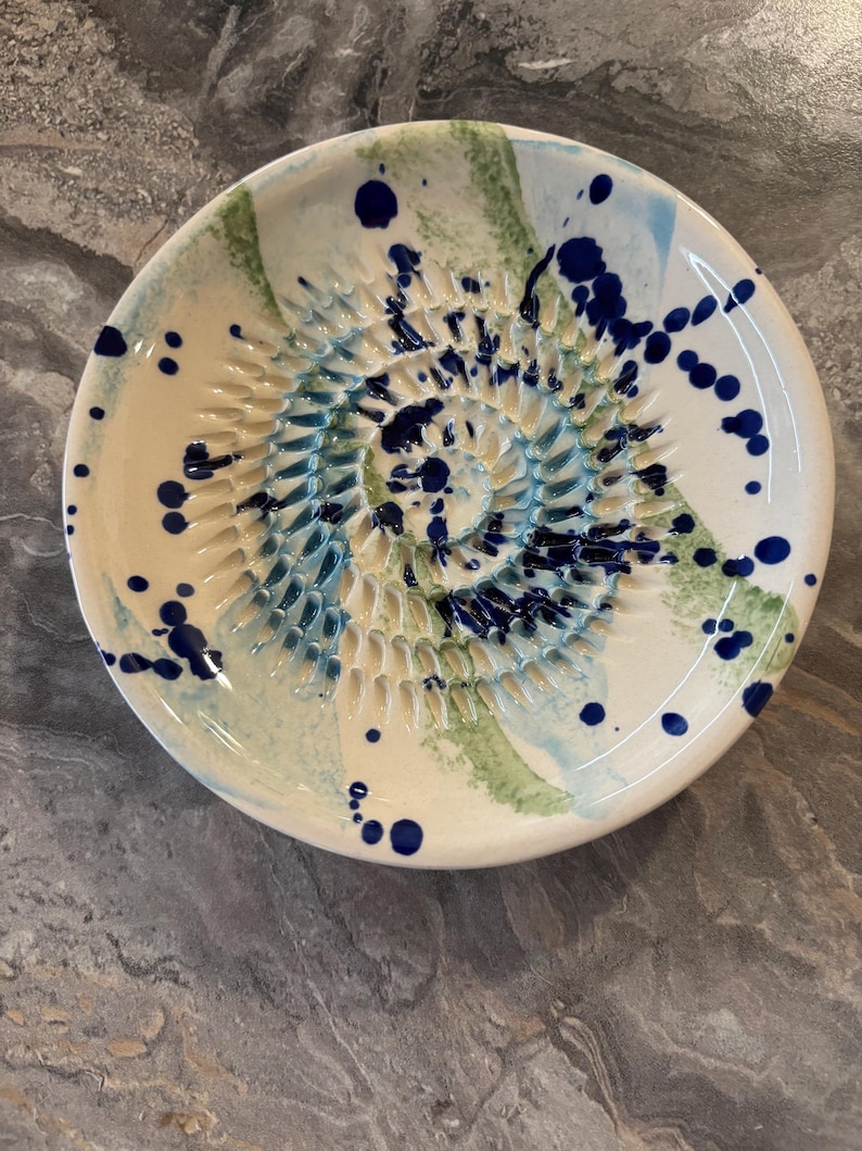 Puede incluir: Un plato rallador de cer&aacute;mica redondo con base color crema, que presenta una espiral central de dientes elevados para rallar. La superficie est&aacute; decorada con salpicaduras y trazos abstractos de pintura azul oscuro, azul claro y verde salvia.